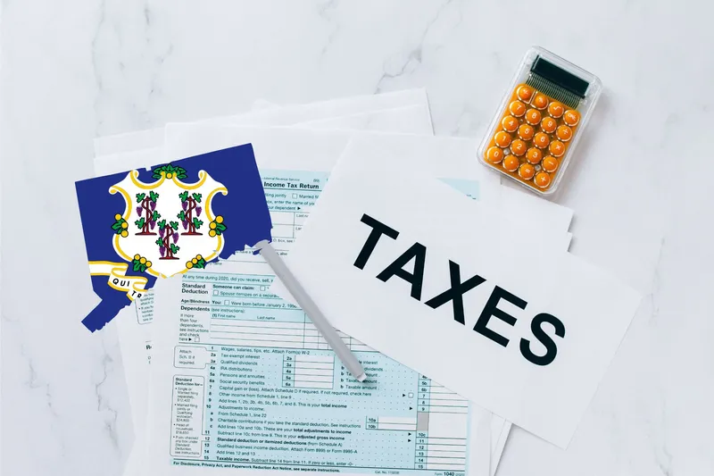 A capital gains calculator sits next to tax forms, a paper with the word 'TAXES', and an Connecticut state logo on a white marble surface.