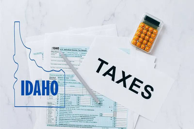 A capital gains calculator sits next to tax forms, a paper with the word 'TAXES', and an Idaho state logo on a white marble surface.