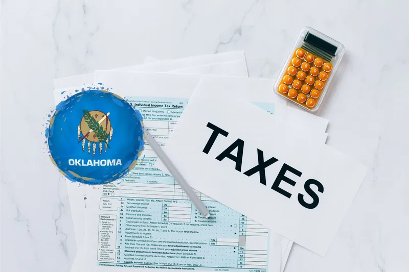 A capital gains calculator sits next to tax forms, a paper with the word 'TAXES', and an Oklahoma state logo on a white marble surface.