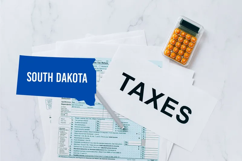 A capital gains calculator sits next to tax forms, a paper with the word 'TAXES', and an South Dakota state logo on a white marble surface.