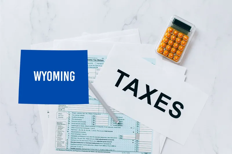 A capital gains calculator sits next to tax forms, a paper with the word 'TAXES', and an Wyoming state logo on a white marble surface.