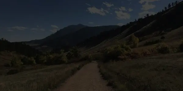 A dirt trail winds through grassy fields and gently rolling Colorado hills, with trees scattered along the path and a mountain rising in the background under a partly cloudy sky.