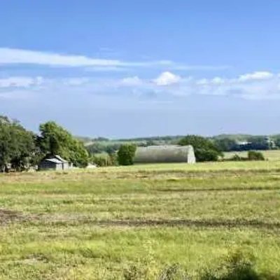 A wide grassy field with scattered trees and a few farm buildings in the background, under a partly cloudy Minnesota blue sky.