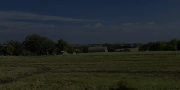 A wide grassy field with scattered trees and a few farm buildings in the background, under a partly cloudy Minnesota blue sky.