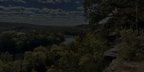 A scenic view of a forested Missouri landscape with a river winding through the trees, seen from a rocky outcrop. The sky is partly cloudy, and the area is lush with greenery.