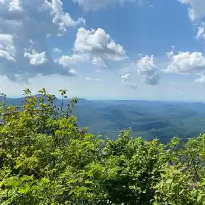 A scenic view from a North Carolina mountaintop with lush green foliage in the foreground and rolling hills stretching into the distance under a partly cloudy sky.