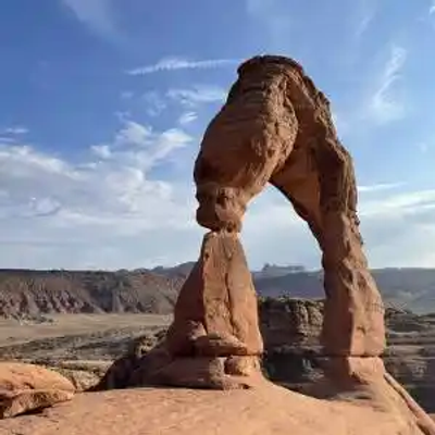 A natural stone arch known as Delicate Arch stands in the Utah desert landscape under a partly cloudy sky, with rocky hills and flat terrain in the background.