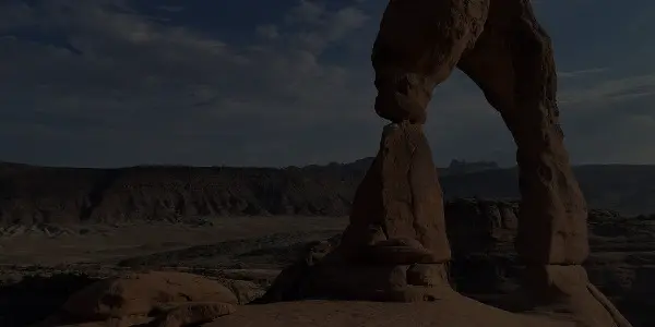 A natural stone arch known as Delicate Arch stands in the Utah desert landscape under a partly cloudy sky, with rocky hills and flat terrain in the background.