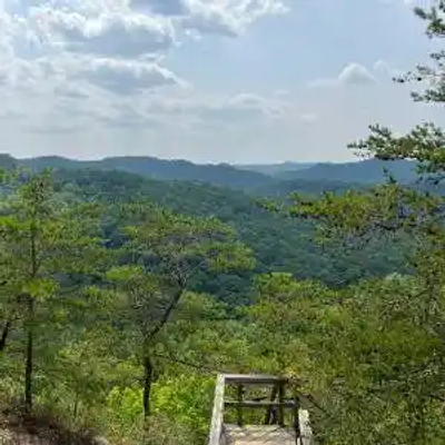 A scenic view from a wooden overlook platform in Kentucky shows a dense forest of green trees with distant rolling hills under a clear sky.