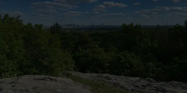 View from a rocky ledge overlooking a dense Massachusetts forest, with a distant city skyline under a sky dotted with clouds.