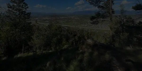 View from a forested hillside in Montana overlooking a highway and a sprawling city, with distant mountains beneath a partly cloudy blue sky.