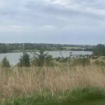 A grassy Nebraska field in the foreground overlooks a lake with boats and trees along the shoreline in the background, under a cloudy sky.