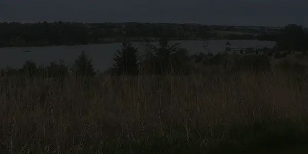 A grassy Nebraska field in the foreground overlooks a lake with boats and trees along the shoreline in the background, under a cloudy sky.