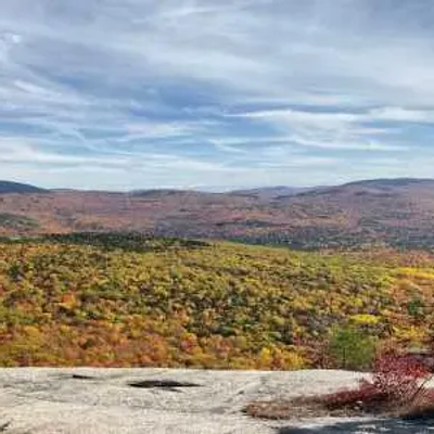 A wide view of a forested New Hampshire valley with autumn foliage in shades of green, yellow, and orange, seen from a rocky overlook under a partly cloudy sky.