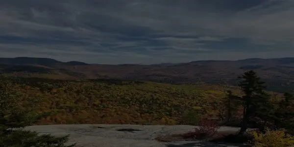 A wide view of a forested New Hampshire valley with autumn foliage in shades of green, yellow, and orange, seen from a rocky overlook under a partly cloudy sky.