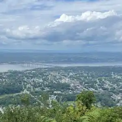 A scenic view from a high vantage point overlooking a river, a large bridge, and a New York town surrounded by green trees and hills under a partly cloudy sky.