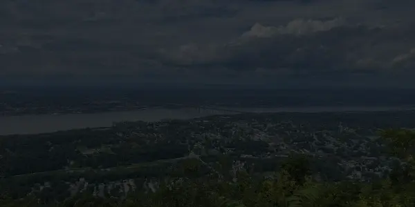 A scenic view from a high vantage point overlooking a river, a large bridge, and a New York town surrounded by green trees and hills under a partly cloudy sky.
