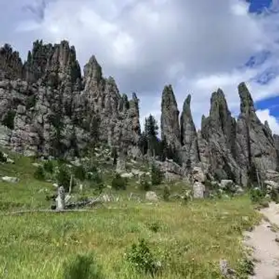 A dirt trail winds through a grassy South Dakota field toward a dramatic rock formation of tall, jagged spires with scattered trees growing among the rocks under a partly cloudy blue sky.